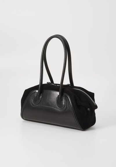Black handbag with long double handles, a leather front panel, and a zipped top, resting on a white background.