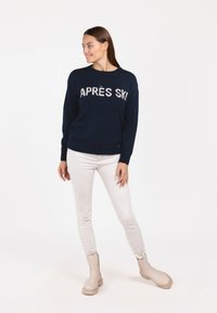 Marineblaue Strickpullover mit dem Text "APRÈS SKI" über der Brust, Rundhalsauschnitt und gerippten Bündchen, kombiniert mit hellen Jeans und beigen Stiefeletten.