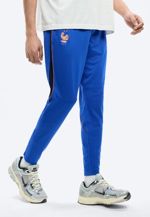 Uomo che indossa pantaloni sportivi blu con rifiniture arancioni e logo della Federazione Francese di Calcio, calzini bianchi e scarpe da ginnastica Nike argento mentre cammina.