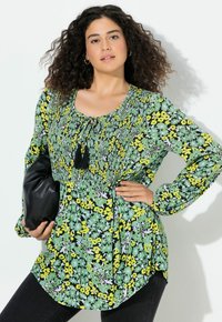 Tunic floreale verde con maniche lunghe, caratterizzata da motivi gialli e bianchi, scollatura arricciata con frange nere e orlo curvo.