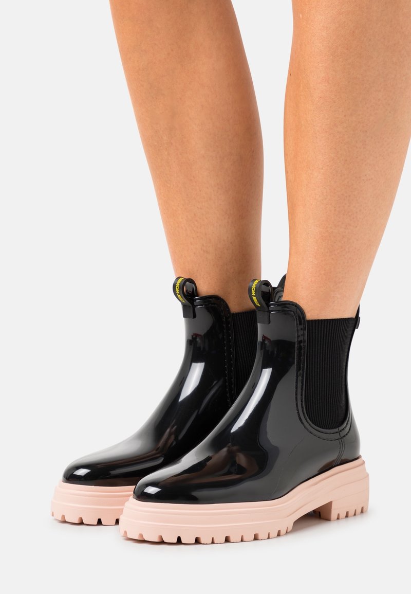 Bottines Chelsea noires en caoutchouc avec une finition brillante, des panneaux latéraux élastiques et une semelle épaisse rose. Présente des tirettes jaunes pour un enfilage facile.