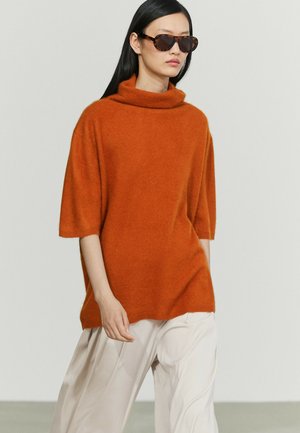 Donna con occhiali da sole tartarugati, maglione oversize arancione bruciato con maniche a metà e pantaloni bianchi larghi su sfondo neutro.