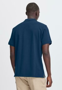 Mann iført en marineblå kortermet polo-skjorte og beige bukser, stående med ryggen til mot en ensfarget lys bakgrunn.