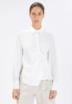 Donna con capelli raccolti che indossa una camicetta bianca a maniche lunghe con dettaglio arricciato ai lati e pantaloni beige con una grande tasca laterale.