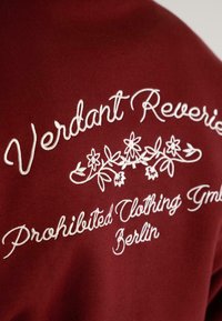 Hvid broderet tekst og blomsterdesign på mørkerød stof, der læser "Verdant Reverie Prohibited Clothing GmbH Berlin."
