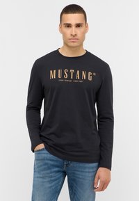 Zwarte langemouwen t-shirt van katoen, met een gouden Mustang-logo op de borst, gecombineerd met blauwe spijkerbroek. Eenvoudig, casual ontwerp.