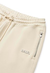 Pantaloni de trening bej cu talie elastică, șnur ajustabil și un buzunar cu fermoar. Prezintă un logo argintiu "BALR." pe partea din față.