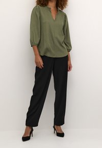 Femme portant un chemisier texturé vert olive avec des manches bouffantes 3/4, un pantalon noir à jambes larges et des escarpins noirs à bout pointu.