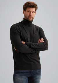 Maglione nero a collo alto in morbido materiale, design aderente, maniche lunghe e una piccola patch con logo sul petto. Abbinato a jeans blu.