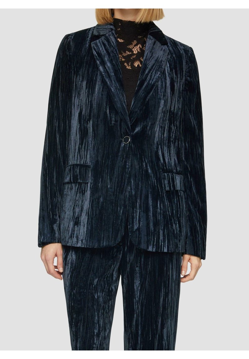 Femme portant un blazer en velours froissé bleu foncé et un pantalon assorti avec un haut en dentelle noire en dessous, debout devant un fond uni.