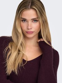 Burgundski rebrasti džemper s V-izrezom, dugim rukavima i mekom teksturom. Kosa je duga i valovita, uokvirujući lice.