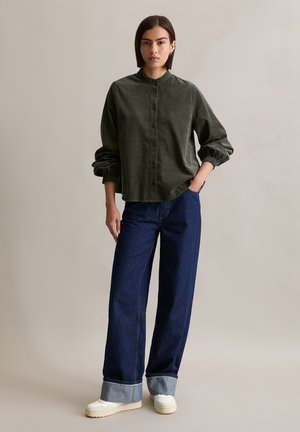 Chemise boutonnée en velours côtelé vert foncé avec un col haut, associée à un jean en denim bleu ample avec des poignets retroussés et des baskets de couleur claire.