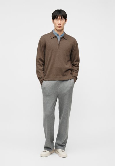 Νεαρός άνδρας που στέκεται με τα χέρια στις τσέπες, φορώντας καφέ πουλόβερ με φερμουάρ στον γιακά, μπλε πουκάμισο, γκρι παντελόνι και λευκά αθλητικά παπούτσια στο λευκό φόντο.