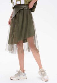 Oliwkowa spódnica z warstwowym, przezroczystym tiulem, asymetrycznym dołem, marszczonym w pasie i srebrnymi akcentami. Połączona z jasnymi trampkami.