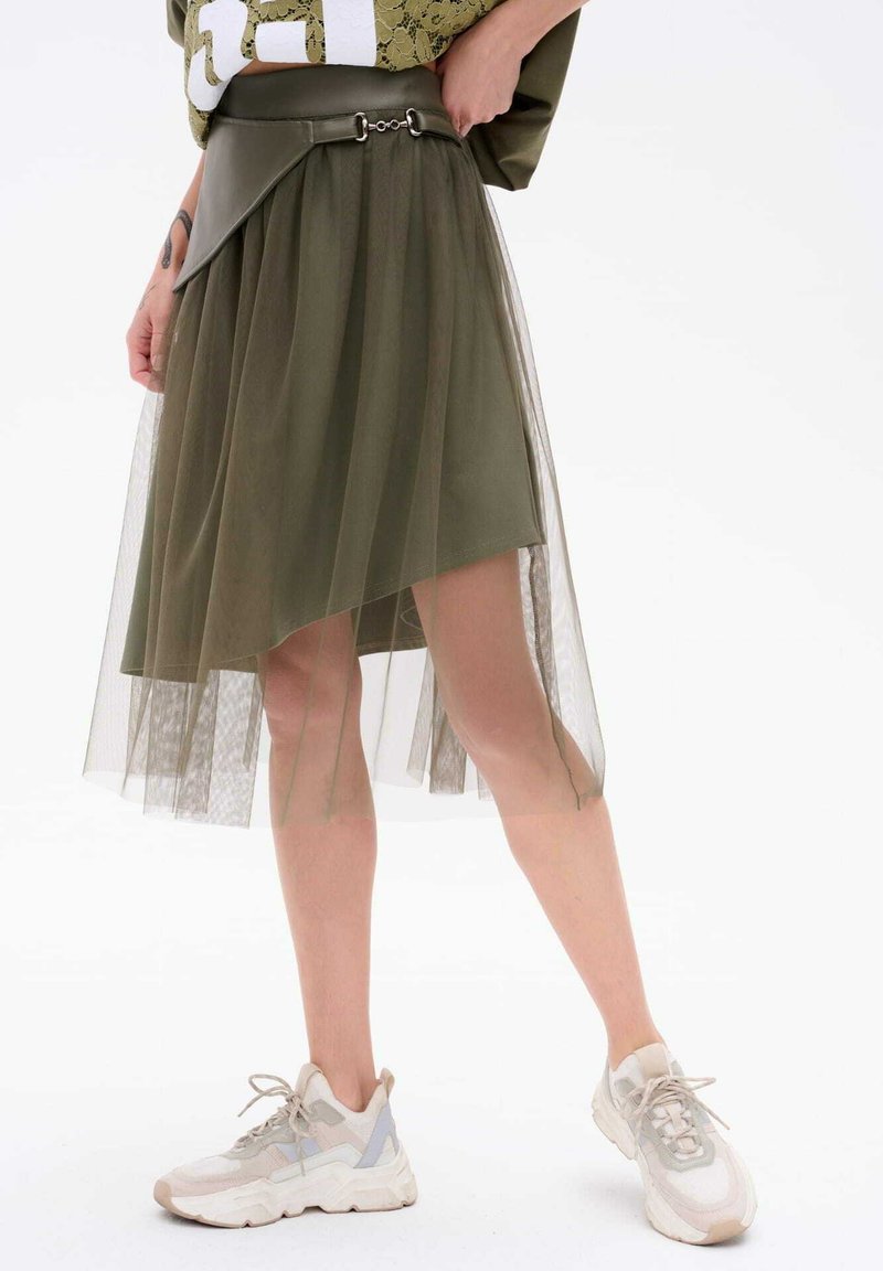 Oliwkowa spódnica z warstwowym, przezroczystym tiulem, asymetrycznym dołem, marszczonym w pasie i srebrnymi akcentami. Połączona z jasnymi trampkami.