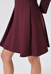 Burgunderfarbenes A-Linie-Kleid mit taillierter Oberseite, gefälteltem Rock und weichem, strukturiertem Stoff. Die Länge reicht bis über das Knie.