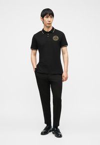 Άνδρας που φοράει μαύρο πόλο με χρυσή διακόσμηση και λογότυπο, μαύρο παντελόνι και μαύρα μοκασίνια, στέκεται με το ένα χέρι στην τσέπη πάνω σε λευκό φόντο.