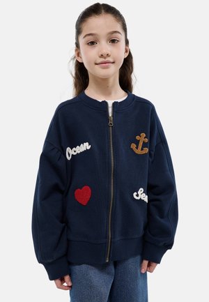 Fille aux longs cheveux bruns portant une veste zippée bleu marine ornée d'une ancre, d'un cœur et des mots « Ocean » et « Sea » sur un jean bleu.