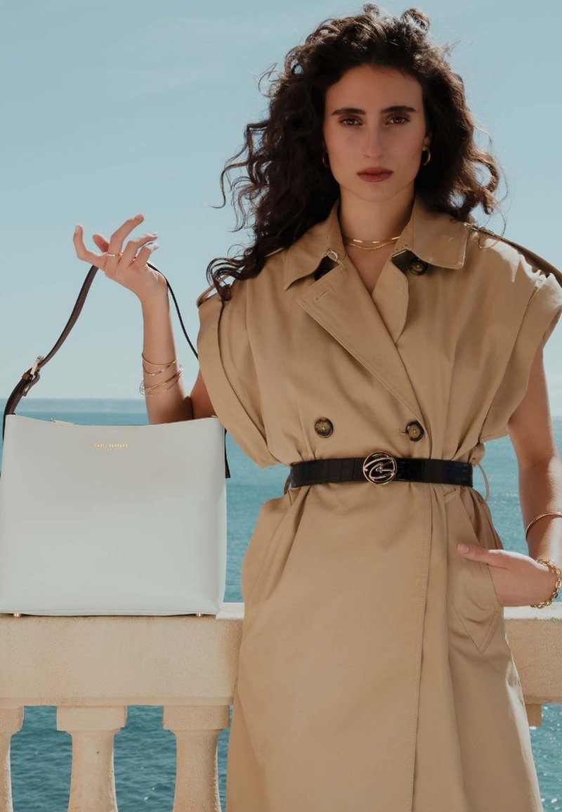 Beige trenchcoat met oversized mouwen, met een riem in de taille. Een witte totebag met een bruine strap wordt vastgehouden in de hand van het model, met een zeed achtergrond.