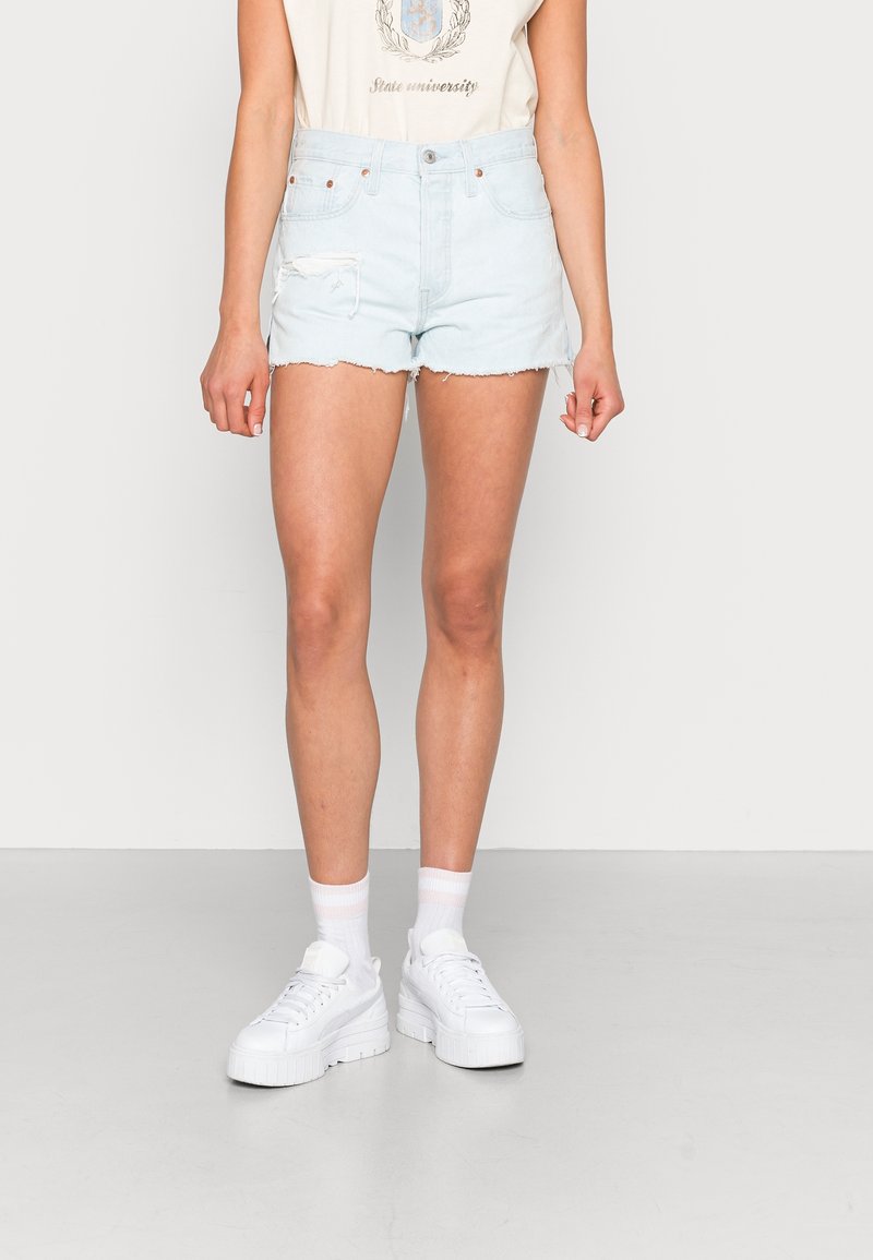 Femme portant un short en jean bleu clair délavé, un t-shirt blanc à motif, des chaussettes blanches et des baskets à semelles compensées blanches, debout devant un fond uni.
