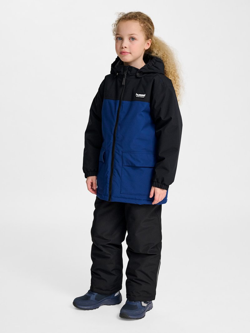 Kind met krullend haar dat een blauw-zwarte capuchonjas, zwarte broek en blauwe sneakers draagt, staand tegen een effen achtergrond.