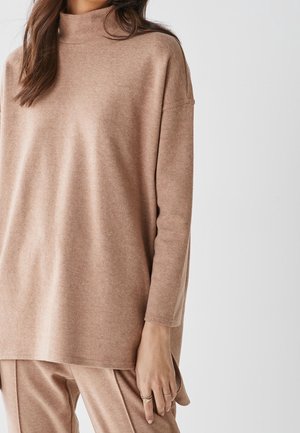 Femme portant un pull à col roulé beige ample et un pantalon assorti, avec des bagues aux doigts et des cheveux bruns sur les épaules.