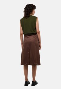 Femme aux cheveux bouclés portant un haut sans manches vert olive, une jupe midi marron avec poches arrière, et des chaussures noires, debout de dos.