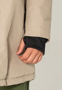 Beige gepolsterte Jacke mit einem schwarzen Ärmelbund. Sichtbare Hand, die den Bund hält. Die Jacke hat eine glatte Textur und ein tailliertes Design.