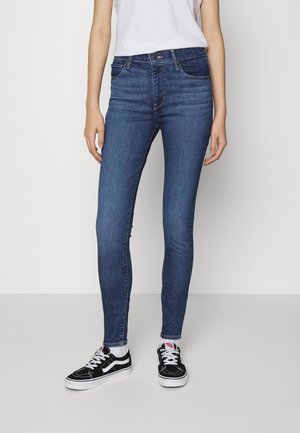Mujer vistiendo jeans skinny azul oscuro, calcetines blancos y zapatillas negro y blanco, de pie contra un fondo gris liso.