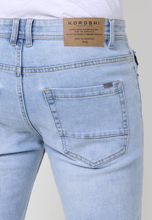 Šviesiai mėlyni džinsai, su siauru siluetu, turintys odinį ženkliuką juosmens srityje ir užpakalinį kišenę su kontrastingomis siūlėmis.