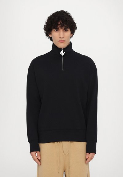 Pulover negru din fleece cu guler înalt și fermoar, dotat cu un tab de tragere cu logo alb, asociat cu pantaloni bej cu șnur. Textură moale, croi lejer.