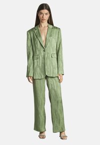 Vrouw in een gemêleerde lichtgroene oversized blazer en bijpassende wijde broek met open teenhakken met kettingband, rechtopstaand.