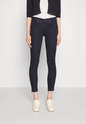 Mujer con vaqueros pitillo azul oscuro, una camiseta blanca con textura, un cárdigan corto negro y mocasines crema, de pie frente a un fondo liso.