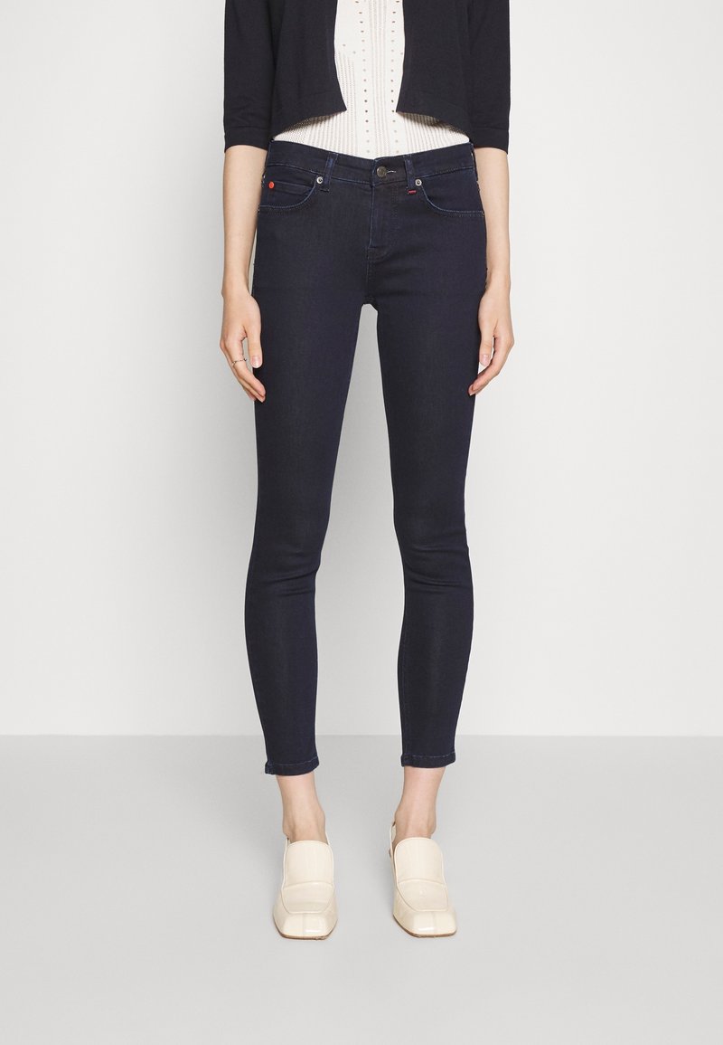 Mujer con vaqueros pitillo azul oscuro, una camiseta blanca con textura, un cárdigan corto negro y mocasines crema, de pie frente a un fondo liso.