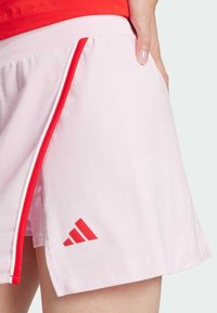 Svetle růžové sportovní kraťasy s červeným pruhem po straně, s malým červeným logem Adidas blízko lemu a elastickým pasem.