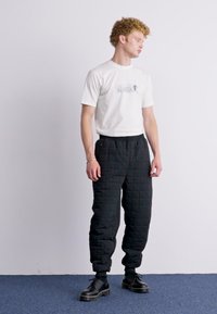 T-shirt blanche avec motif imprimé, pantalons de survêtement matelassés noirs et bottes à lacets noires ; le modèle se tient contre un fond uni.