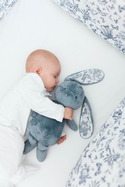 Μαλακό μπλε κουνελάκι παιχνιδιού με μεγάλες σαν αυτιά και εσωτερικό ύφασμα με ανθισμένο μοτίβο. Βρέφος ντυμένο στα λευκά κοιμάται δίπλα του σε λευκό σεντόνι με μπλε ανθισμένο κλινοσκεπάσμα.