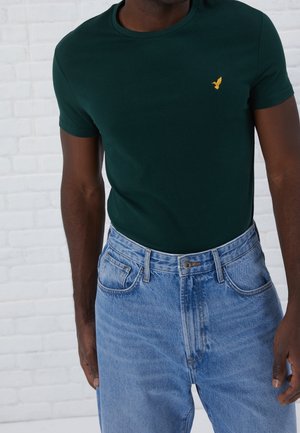 Homme portant un t-shirt vert foncé ajusté avec un petit logo d’oiseau jaune et un jean taille haute bleu clair devant un mur de briques blanches.