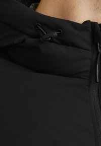 Musthave musta tõmblukuga jakk, millel on kõrge krae, sile kangas, reguleeritavad nöörid ja detailne õmblus tõmbluku lähedal.