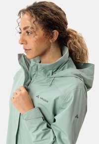 Giacca impermeabile verde chiaro con colletto alto, cappuccio regolabile e tasche con zip. Presenta il logo sul petto e un tessuto testurizzato.