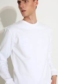 Bijela dukserica s okruglim izrezom, izrađena od mekog pamuka, s dugim rukavima i rebrastim manžetama, koja ima jednostavan, minimalistički dizajn.