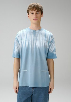 Ung mand iført en løs lyseblå tie-dye t-shirt med hvid tekst, blå jeans, stående imod en ensfarvet grå baggrund.