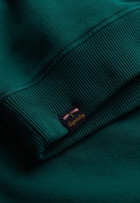 Felpa verde con polsini e orlo a coste, morbida al tatto, con un piccolo logo ricamato di Superdry e il dettaglio di due bandiere.