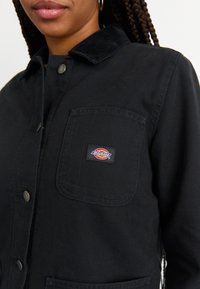 Melns kanvas žakets ar kordurojas apkakli, metāla pogām un krūškurvja kabatu, kurā ir daudzkrāsains Dickies logo.