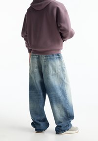 Jeans in denim sbiadito a gamba larga con una tonalità di blu chiaro; presentano due tasche posteriori. Indossati con una felpa con cappuccio scura e sneakers grigie.