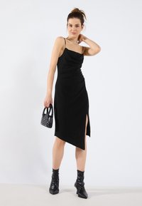Robe noire sans manches avec un col drapé et un ourlet asymétrique. Présente une fente sur le côté et un tissu texturé, associée à un petit sac à main clouté.