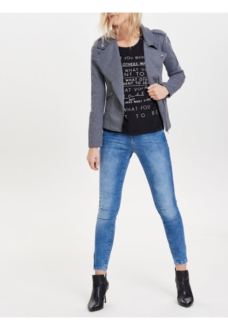 Femme portant une veste grise à fermeture éclair, un t-shirt noir imprimé, un jean skinny bleu et des bottines noires, debout les jambes écartées devant un fond blanc.