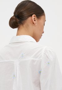 Bluză albă cu guler, decorată cu broderie delicată în flori de margaretă albastre și detalii pliate pe umeri. Material moale, ușor.