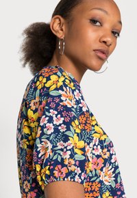 Blusa floral com fundo azul-marinho; apresenta flores vibrantes em amarelo, rosa e branco. Mangas curtas com volume e textura de tecido suave.