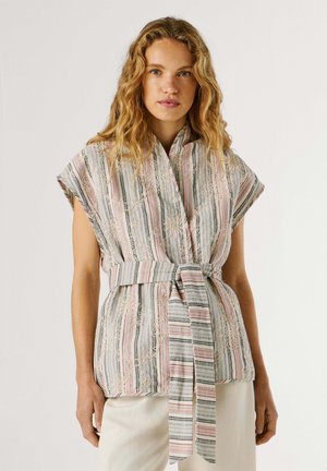 Donna con capelli biondi ondulati che indossa un gilet senza maniche a righe con ricami floreali e una cintura coordinata annodata in vita.